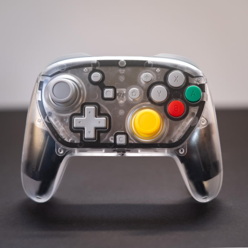 May include: A transparent video game controller with a clear plastic shell. The controller features a gray joystick, a yellow center button, and colored buttons in green, red, and gray. The design includes a directional pad and various function buttons.