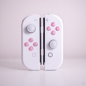 Pode incluir: Controlador de jogos branco com botões rosa e joysticks cinzentos. O controlador é dividido em duas partes, com os botões e joysticks na frente. Os botões rosa têm um efeito marmoreado. Projetado para um console de jogos.