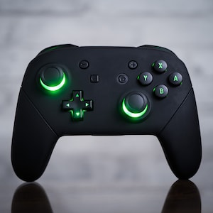 May include: A black video game controller with green glowing buttons. The controller has a directional pad, four face buttons, two shoulder buttons, and two analog sticks.