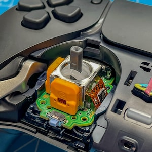 May include: Close-up of a disassembled video game controller, showing the internal components and circuitry. The joystick is visible, along with a small circuit board and a grey plastic component.