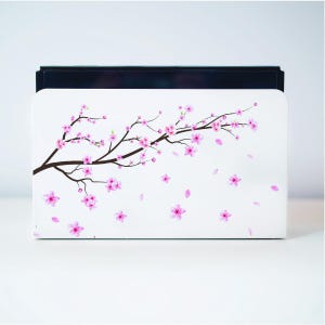 May include: White Nintendo Switch game case with a pink cherry blossom branch design. The branch has pink flowers and petals falling from it.