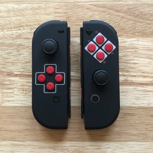 May include: A black and red Nintendo Switch controller with a retro-style design. The controller has a directional pad, four face buttons, and two shoulder buttons. The face buttons are labeled "A", "B", "X", and "Y".