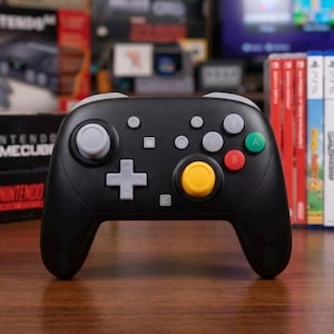 May include: A black wireless game controller featuring a light gray directional pad, a yellow right analog stick, and a light gray left analog stick. The face buttons include a green A button, a red B button, a gray X button, and a gray Y button.