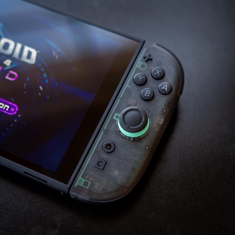 Puede incluir: Primer plano de un mando de consola de videojuegos negro y transparente. El mando tiene una pantalla que muestra el t&iacute;tulo de un juego. Los botones son negros, con un joystick verde brillante. El mando est&aacute; sobre una superficie negra.