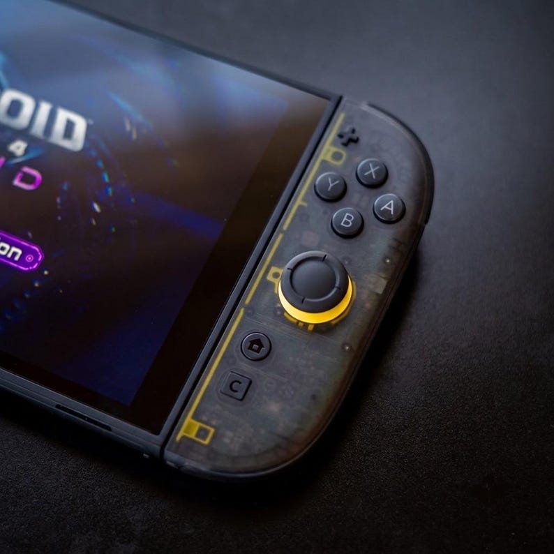 Puede incluir: Una consola de juegos port&aacute;til con un mando de color gris oscuro transl&uacute;cido que deja ver sus componentes internos. El mando incluye botones negros A, B, X e Y, una cruceta y un joystick central con un anillo amarillo brillante.