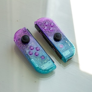 May include: Two iridescent blue and purple Nintendo Switch Joy-Con controllers with glitter accents. The controllers have black buttons and a black analog stick.