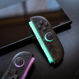 Puede incluir: Primer plano de dos mandos de juego negros transparentes y verde menta con botones negros. Uno de los mandos est&aacute; inclinado hacia el espectador, el otro est&aacute; en la esquina inferior izquierda. Los mandos est&aacute;n sobre una superficie negra.