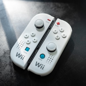 Puede incluir: Dos mandos remotos Wii blancos con detalles en negro. Cada mando tiene un joystick, botones direccionales y botones de acción. Los mandos están etiquetados con el logotipo de Wii y tienen un botón de encendido y un botón de inicio.