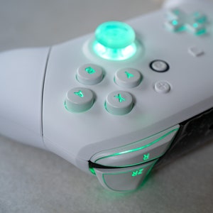 May include: A white gaming controller with green illuminated buttons, including the A, B, X, and Y buttons, as well as the directional pad and the right and left triggers.