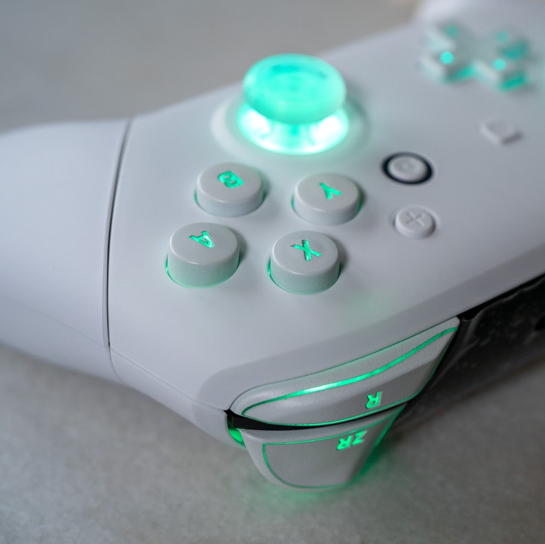 May include: A white gaming controller with green illuminated buttons, including the A, B, X, and Y buttons, as well as the directional pad and the right and left triggers.
