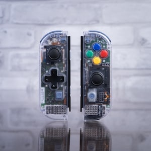 May include: A pair of clear, transparent Nintendo Switch Joy-Con controllers with black buttons. The controllers have a translucent design, revealing the internal components.