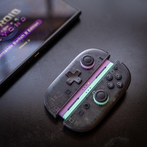 Puede incluir: Un mando transparente negro de Nintendo Switch con detalles en morado y verde. El mando tiene una cruceta direccional, botones y joysticks. El texto "Nintendo Switch 2 Edition" es visible en una pantalla en el fondo.