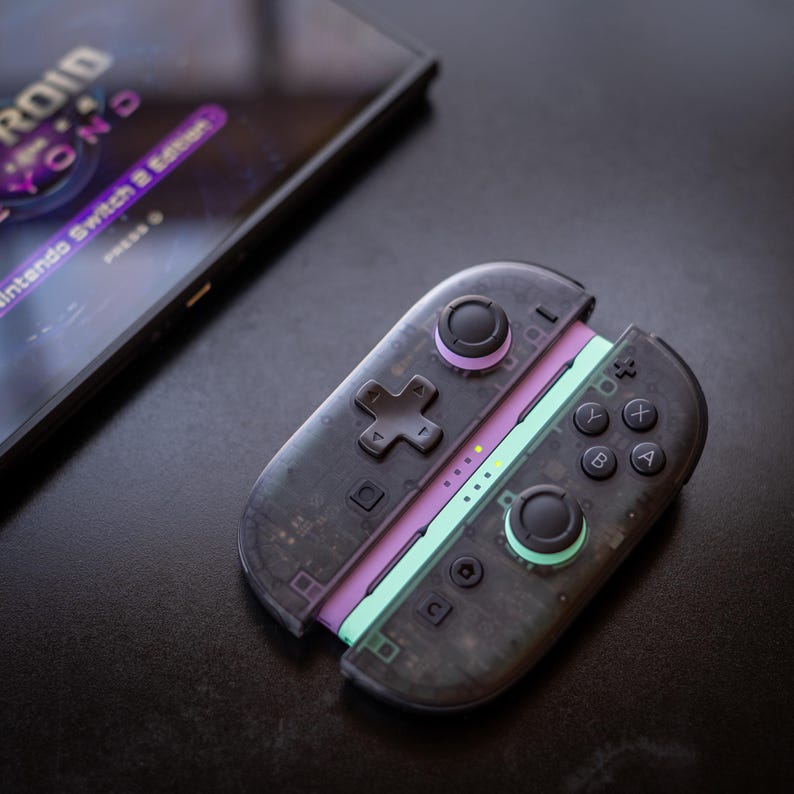 Puede incluir: Un mando transparente negro de Nintendo Switch con detalles en morado y verde. El mando tiene una cruceta direccional, botones y joysticks. El texto "Nintendo Switch 2 Edition" es visible en una pantalla en el fondo.