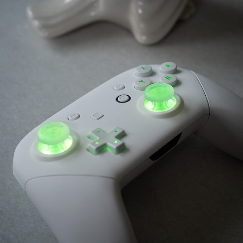 May include: A white gaming controller with green glowing buttons. The controller has a directional pad, four face buttons, and two analog sticks.