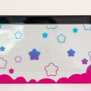 Pode incluir: Console Nintendo Switch branca com uma pele personalizada rosa e branca com um padr&atilde;o de nuvens e estrelas. A pele tem um degradado de estrelas azuis e roxas.