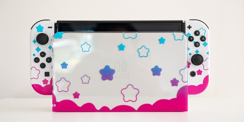 Pode incluir: Console Nintendo Switch branca com uma pele personalizada rosa e branca com um padr&atilde;o de nuvens e estrelas. A pele tem um degradado de estrelas azuis e roxas.