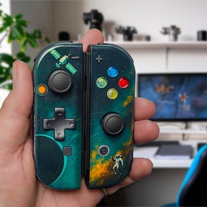 May include: A pair of teal and black video game controllers with a space theme. One controller features a satellite graphic, while the other has an astronaut and colorful buttons. The controllers are held in a person's hand.