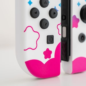 Pode incluir: Um controlador Nintendo Switch Joy-Con branco e rosa com um design colorido com estrelas, nuvens e flores. O controlador tem um bot&atilde;o rosa e um bot&atilde;o preto.