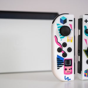 May include: A pair of white Nintendo Switch Joy-Con controllers with a retro 80s design. The controllers feature a variety of colorful geometric shapes, including triangles, squares, and circles. The left controller has a pink Game Boy-style design, while the right controller has a colorful Rubik's Cube design.