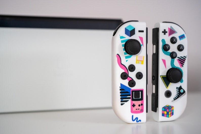 May include: A pair of white Nintendo Switch Joy-Con controllers with a retro 80s design. The controllers feature a variety of colorful geometric shapes, including triangles, squares, and circles. The left controller has a pink Game Boy-style design, while the right controller has a colorful Rubik's Cube design.
