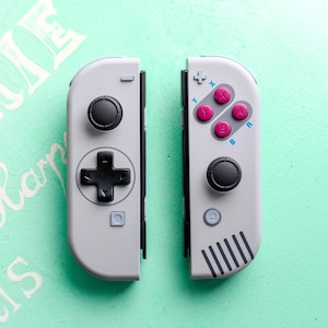 Może przedstawiać: Dwa szare i czarne kontrolery Nintendo Switch z biało-czarnym wzorem. Kontrolery mają krzyżak, cztery przyciski i dwa przyciski na ramionach. Kontrolery znajdują się na jasnozielonej powierzchni.