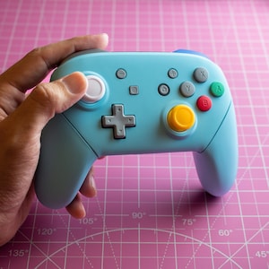 Può includere: Un controller di gioco wireless blu chiaro con un pulsante bianco, un pulsante giallo e pulsanti colorati etichettati X, Y, A e B.