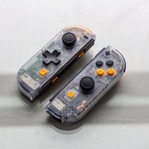 May include: Two clear plastic Nintendo Switch controllers with black buttons and yellow accents. The top controller has a D-pad and the bottom controller has a cross button.