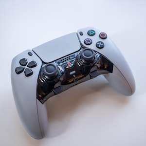 Peut inclure: Une manette de jeu grise et noire avec des joysticks, des boutons et un pavé directionnel noirs. La manette a une section centrale noire brillante avec le logo PlayStation et des boutons d'action colorés.