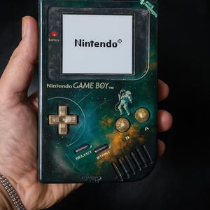 Puede incluir: Una consola portátil Nintendo Game Boy vintage, con un diseño personalizado con temática espacial. La pantalla muestra el logotipo de Nintendo. El dispositivo tiene una cruceta, botones A y B, y botones Select y Start. El Game Boy se sostiene en una mano.
