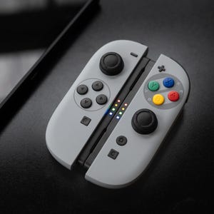 Pode incluir: Um comando de videogame cinza claro com joysticks e botões pretos. O comando tem botões coloridos e luzes indicadoras. O comando está aberto e repousa sobre uma superfície preta.