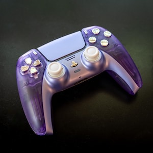 Peut inclure: Une manette de jeu violette transparente avec une section centrale violet clair et des boutons argentés. La manette a un design symétrique avec deux joysticks analogiques, une croix directionnelle et divers boutons.