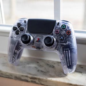 Puede incluir: Un mando de videojuego inalámbrico transparente con panel táctil negro, joysticks negros y botones direccionales negros. El mando incluye los icónicos símbolos de acción en azul, rosa, verde y rojo y un logotipo central de PlayStation.