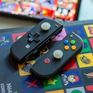 Op de afbeelding: Twee zwarte Nintendo Switch Joy-Con controllers met verschillende knopconfiguraties. Een controller heeft een witte knop en een kruisvormige knop, terwijl de andere een witte knop en kleurrijke ronde knoppen heeft.