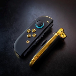 以下が含まれることがあります： 黒と金のゲームコントローラー。 コントローラーは、金色のアクセントとボタンが付いた黒いベースです。 ジョイスティックの周りには青いリングがあります。 コントローラーはゲーム機用に設計されています。