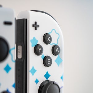 Pode incluir: Um controlador branco do Nintendo Switch com padr&otilde;es geom&eacute;tricos azuis e brancos. O controlador tem bot&otilde;es pretos com as letras X, Y, A e B. O controlador tamb&eacute;m tem um joystick preto.