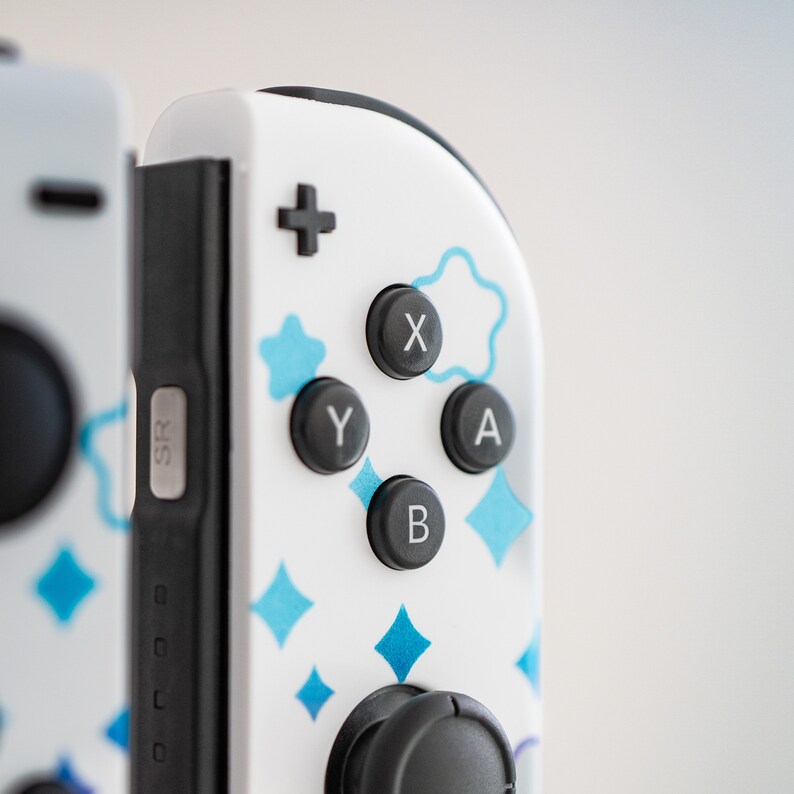Pode incluir: Um controlador branco do Nintendo Switch com padr&otilde;es geom&eacute;tricos azuis e brancos. O controlador tem bot&otilde;es pretos com as letras X, Y, A e B. O controlador tamb&eacute;m tem um joystick preto.