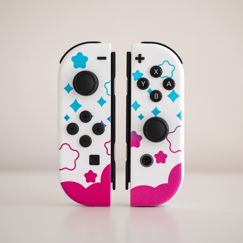 Pode incluir: Um par de controladores Nintendo Switch Joy-Con brancos com um design colorido que inclui estrelas azuis, nuvens cor-de-rosa e as letras "X", "Y", "A" e "B".
