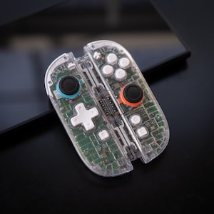 May include: Clear, transparent video game controllers with visible internal components. The controllers have black joysticks with blue and orange accents, white buttons, and a white directional pad. The controllers are resting on a black surface.