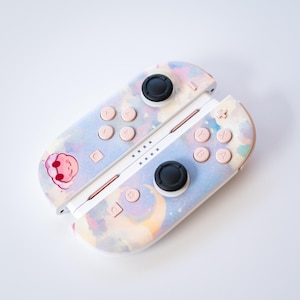 May include: A pair of pastel-colored video game controllers with a dreamy, watercolor-like design. The controllers feature pink buttons, black joysticks, and a small cartoon character. The design includes a crescent moon and stars.