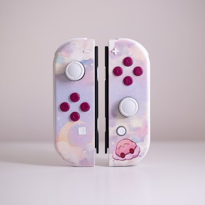 Op de afbeelding: Een witte Nintendo Switch Joy-Con-controller met een roze en paarse aquarelontwerp. De controller heeft een roze Kirby-personage aan de rechterkant.