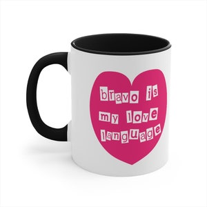 May include: White ceramic coffee mug with a black rim and handle. The mug features a pink heart with the text "bravo is my love language" in white letters.