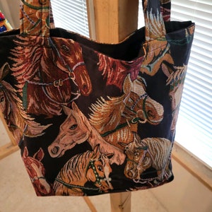 May include: A black tote bag featuring a woven tapestry pattern of multiple horse heads in shades of brown, tan, and white. The bag includes two matching fabric shoulder straps and a deep, open main compartment with a dark interior lining.