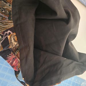 May include: A tote bag featuring a woven tapestry exterior with a multi-colored animal pattern and a solid black fabric lining. The bag is open, revealing the dark interior and the detailed texture of the woven outer material with visible stitching.