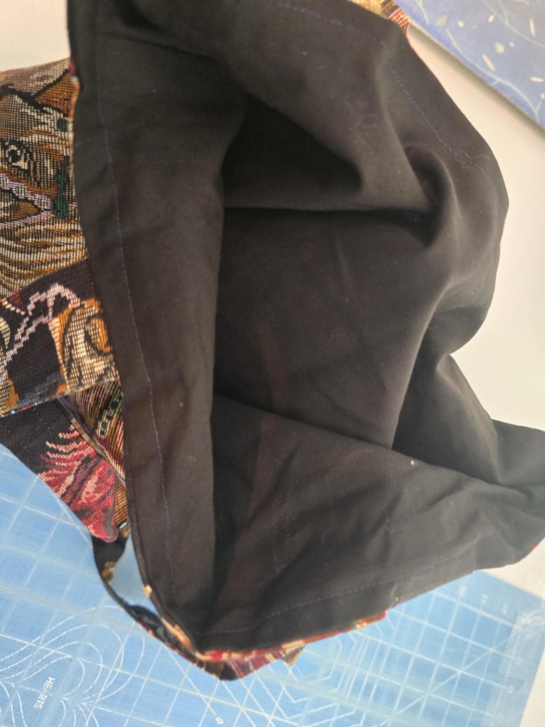 May include: A tote bag featuring a woven tapestry exterior with a multi-colored animal pattern and a solid black fabric lining. The bag is open, revealing the dark interior and the detailed texture of the woven outer material with visible stitching.