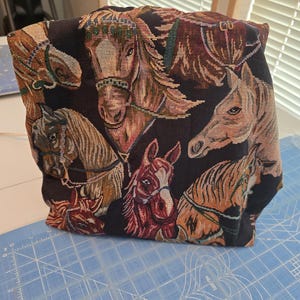 May include: A fabric pouch featuring a woven tapestry design of multiple horse heads in shades of brown, tan, and black against a dark background. The horses are depicted with various bridles and manes, creating a detailed, textured surface.