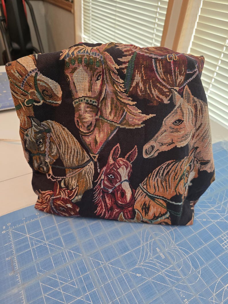 May include: A fabric pouch featuring a woven tapestry design of multiple horse heads in shades of brown, tan, and black against a dark background. The horses are depicted with various bridles and manes, creating a detailed, textured surface.