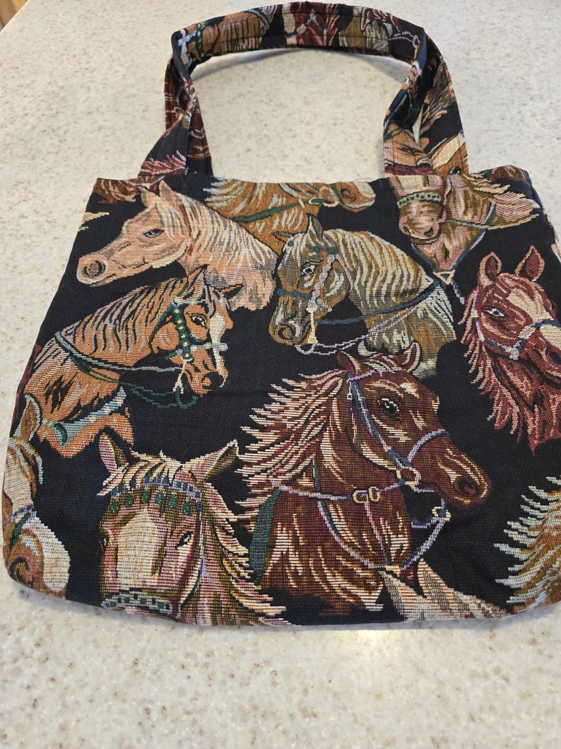 May include: A black tote bag featuring a woven tapestry pattern of multiple horse heads in shades of brown, tan, and white. The bag has two matching fabric handles and a rectangular shape, showcasing detailed equine portraits across the entire surface.