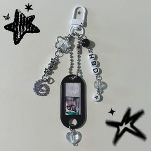 May include: A black keychain with a decorative design. The keychain features a black iPod-shaped charm with a photo inside, a clear heart charm, and beads spelling "HBD". Additional charms include a star and a flower.