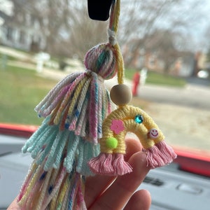 May include: A yellow rainbow-shaped yarn ornament with pink, blue, and green yarn tassels. The rainbow has a green flower, a blue gem, and a yellow smiley face.