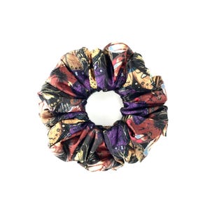Op de afbeelding: Een zwarte scrunchie met een kleurrijke vlinderprint. De print heeft tinten van paars, rood, goud en blauw.